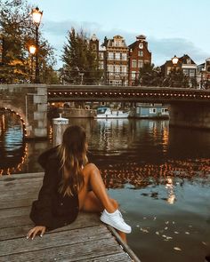 a woman sitting on a dock next to the water with buildings in the background at dusk