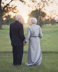 an older couple holding hands while standing in a field with trees and grass at sunset