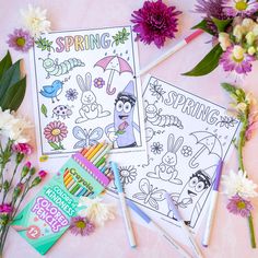 two coloring books and some flowers on a pink surface with markers, pencils and crayons