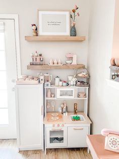 a kitchen with white cabinets and pink accessories on the counter top, next to a wooden shelf filled with dishes