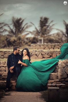 a man and woman posing for a photo in front of stone wall with palm trees