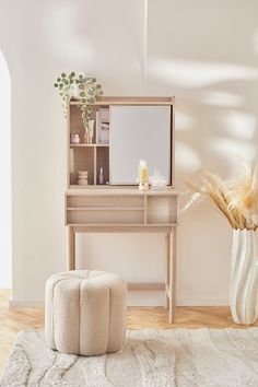 a white vase with dried plants and a mirror on top of a wooden table next to a stool