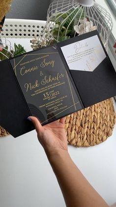 a person holding up an open black and gold wedding card with the wording on it