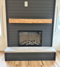 an empty fireplace in a living room with wood flooring and gray walls, along with two windows