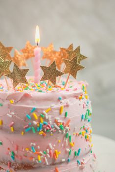 a pink birthday cake with sprinkles and stars on top is lit by a single candle