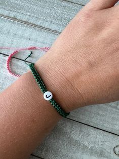 a person wearing a green bracelet with a white beaded initial on the clasp and a pink string