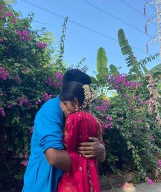 two women hugging each other in front of some bushes and flowers on a sunny day