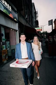 a man and woman are walking down the street holding a pizza box in their hands