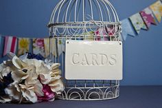 a white birdcage with cards on it next to flowers