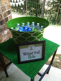 a large green bucket filled with water bottles on top of a grass covered table next to a sign that says watering hole