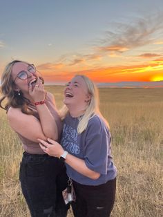 two women are laughing while standing in a field with the sun going down behind them