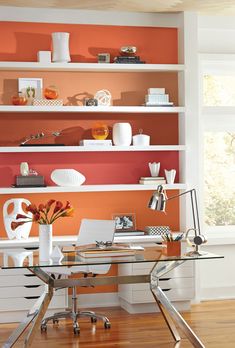 an office with orange walls and white shelvings on the wall, along with a glass top desk