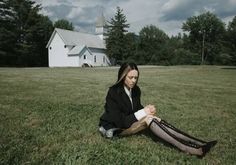 a woman sitting on the grass in front of a church holding her purse and looking down