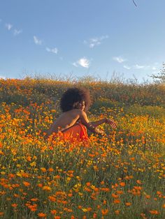 a woman in an orange dress sitting in a field of wildflowers with a kite flying overhead