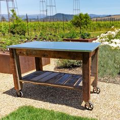 a wooden table sitting in the middle of a gravel ground next to flowers and plants