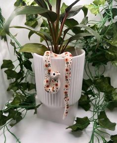 a plant in a white vase with an animal decoration on the front and bottom, surrounded by greenery