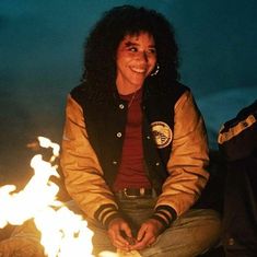 a woman sitting on the ground next to a fire with her hands in her pockets