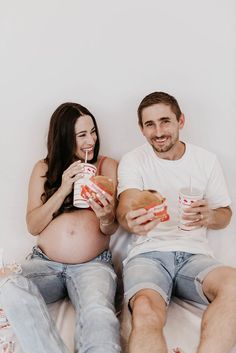 a pregnant couple sitting on a bed eating sandwiches and drinking sodas