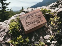 a wooden box that says the adventure begins on top of some rocks and plants with mountains in the background