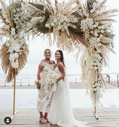 two women standing in front of an arch with white flowers and greenery on it