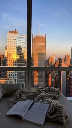 an open book on a bed in front of a window with the city skyline outside