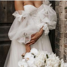 a woman in a white wedding dress holding flowers next to a brick wall and door