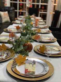 the table is set for thanksgiving dinner with gold plates and silverware, pumpkins and greenery