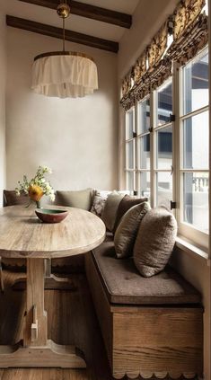 a wooden table sitting in front of a window next to a long bench with pillows on it