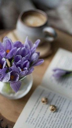 purple flowers sit in a vase on a table next to an open book and cup of coffee