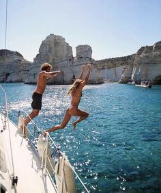 two people jumping off the side of a sailboat into the blue water with cliffs in the background