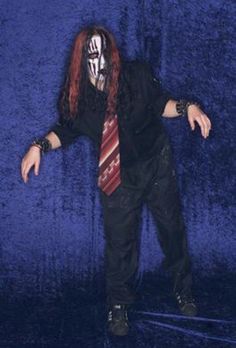 a man with long red hair wearing a black shirt and tie, standing in front of a blue background