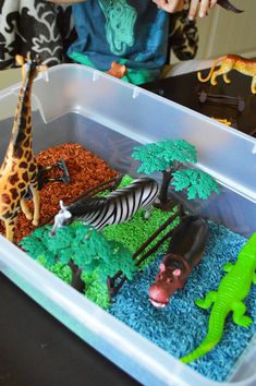 a plastic container filled with fake animals and plants on top of a table next to a child's hand