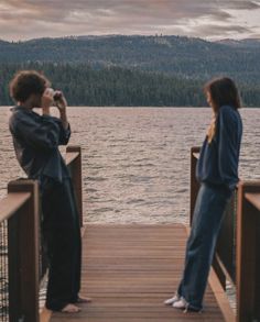 two people are standing on a dock looking at the water and mountains in the distance