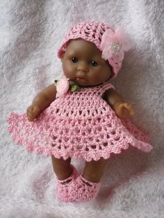 a baby doll wearing a pink crocheted dress and booties on a white blanket