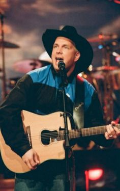 a man wearing a cowboy hat and holding a guitar in front of a microphone on stage