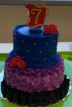 there is a blue and purple cake with red roses on the bottom, and a number seven on top