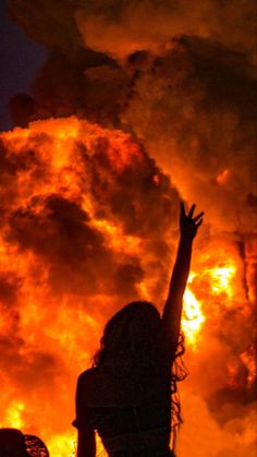 a woman reaching up to the sky with her arms raised in front of a huge fire