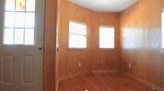 an empty room with wood paneling and two windows in the wall, along with a door to another room