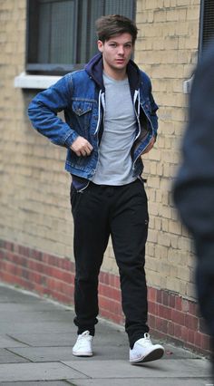 a young man is walking down the sidewalk in front of a brick building wearing a denim jacket and white sneakers