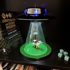 a toy cow under a glass dome on top of a book shelf next to dice