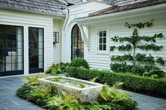 a white house with a fountain in the middle of it's yard and lots of greenery