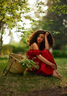 a woman in a red dress sitting on the ground next to a basket and tree