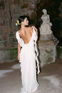 a woman in a white dress standing next to a stone wall with a flower in her hair