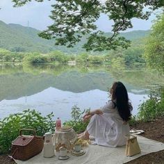 a woman sitting on top of a blanket next to a lake with a tea pot