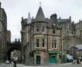 an old stone building with a gate in front of it and people walking by on the sidewalk