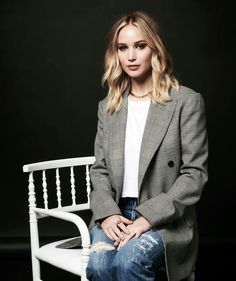 a woman sitting on top of a white chair in front of a black background wearing jeans and a blazer