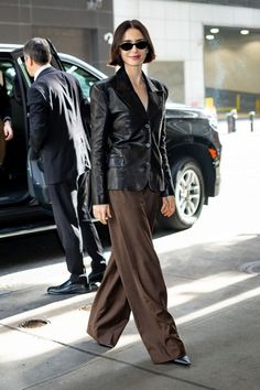 a woman in black jacket and brown pants walking down the street with her hand on her hip