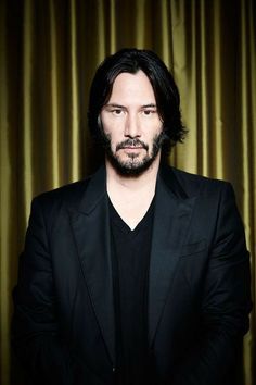 a man with long hair and beard standing in front of a curtained background wearing a black suit