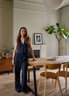 a woman standing next to a wooden table in a room with chairs and a potted plant