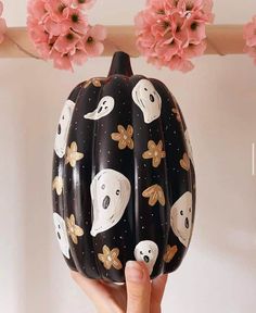 a hand holding up a painted pumpkin with ghost faces on it and flowers in the background